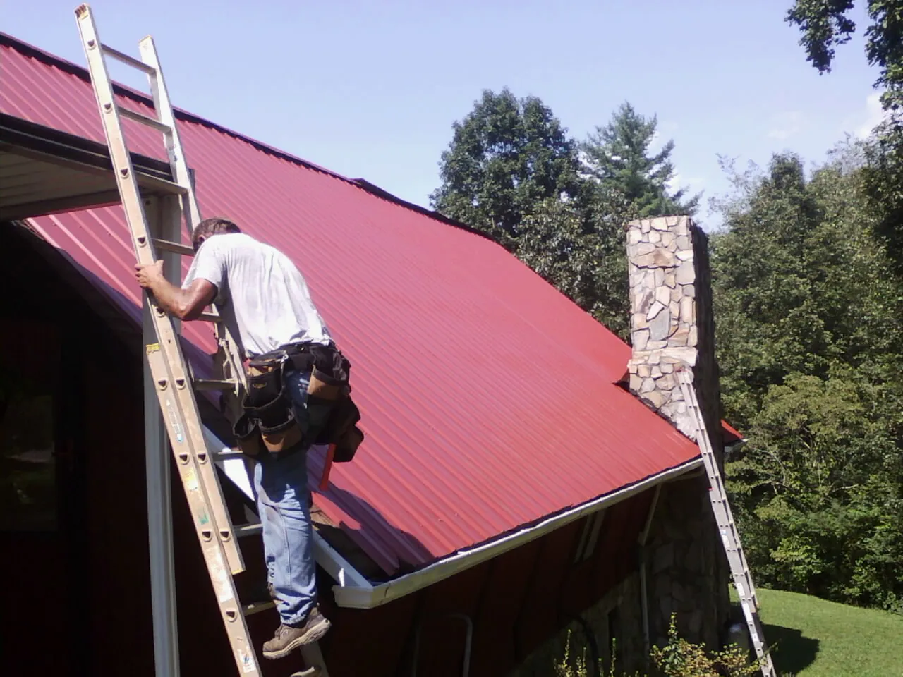Roofer on a ladder beside a red metal roof and stone chimney — Soffit & Fascia Repair by Roofing Brothers in Tyler Hill